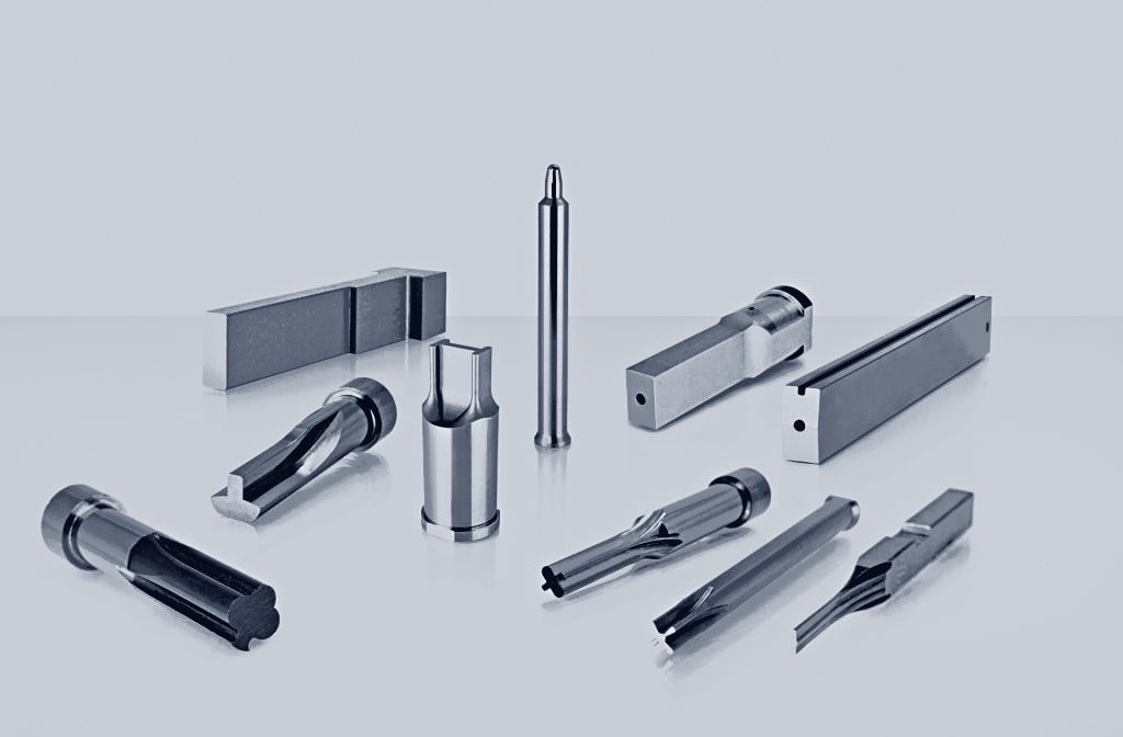 A set of precision punches, guide pins and tooling blades in polished and blackened steel, laid out on a white background.